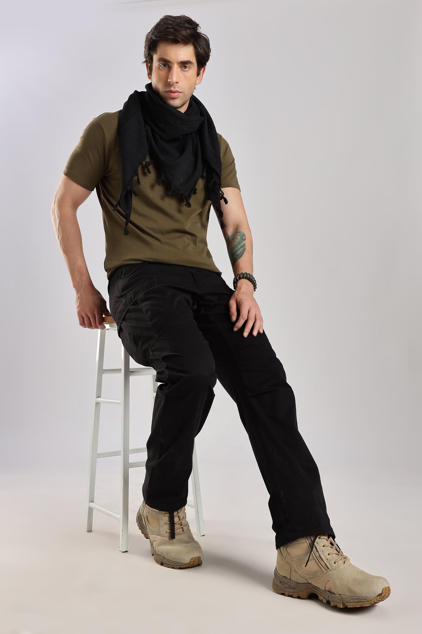 Man sitting on a stool wearing a black tactical shemagh scarf, an olive green tshirt, black cargo pants, and desert combat boots with a plain background.