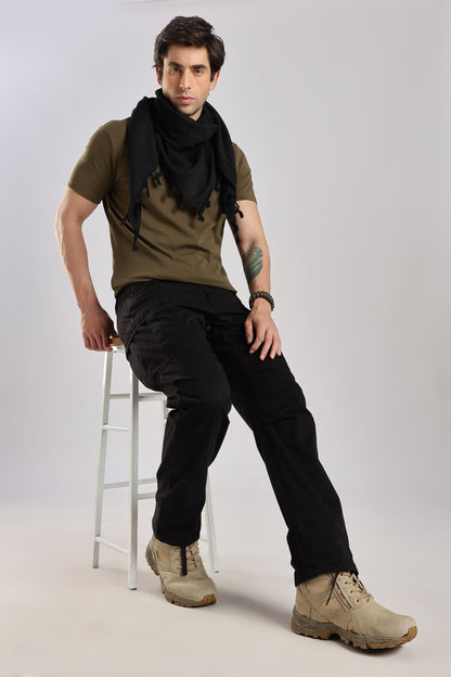 Man sitting on a stool wearing a black tactical shemagh scarf, an olive green tshirt, black cargo pants, and desert combat boots with a plain background.