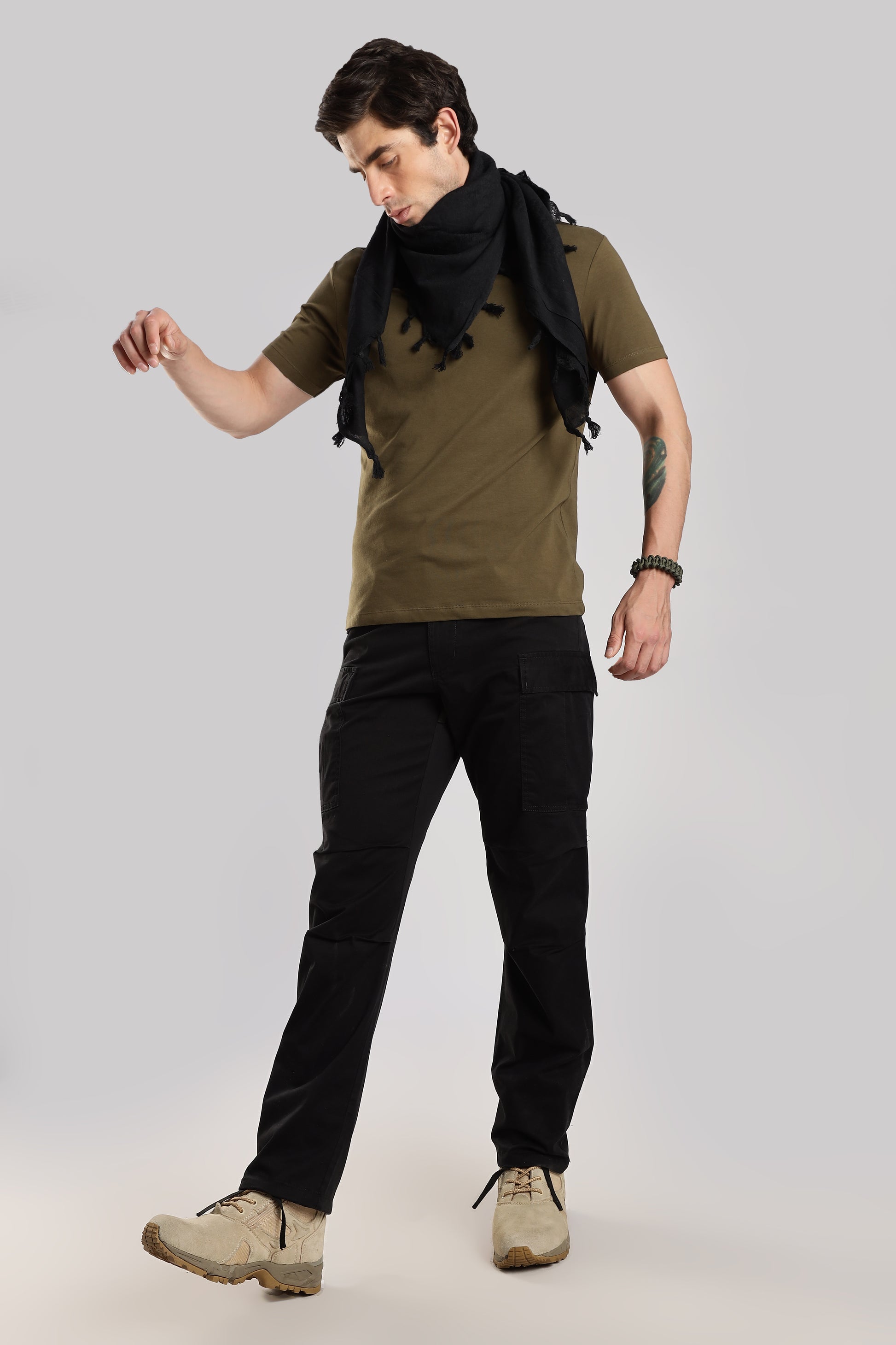 Man wearing a black tactical shemagh scarf, an olive green tshirt, black cargo pants and desert combat boots on a white background.