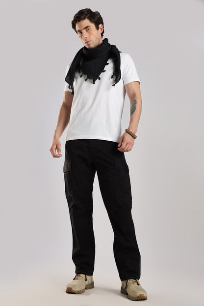 Man wearing a black tactical shemagh scarf, a white t-shirt and a black cargo pants on a grey background.