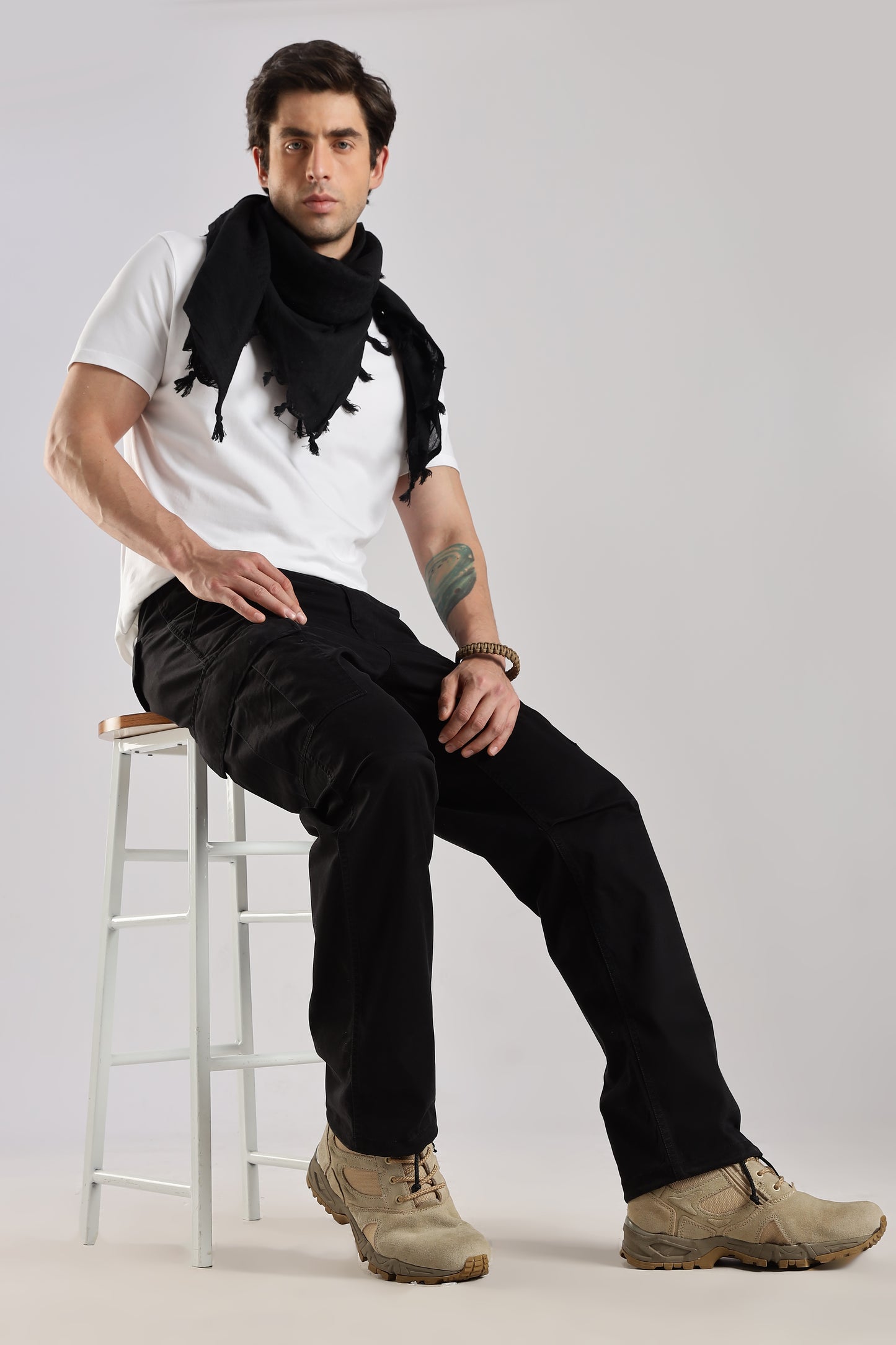 Man sitting on a stool wearing a black tactical shemagh scarf, a white t-shirt, black cargo pants, and desert combat boots against a plain background.