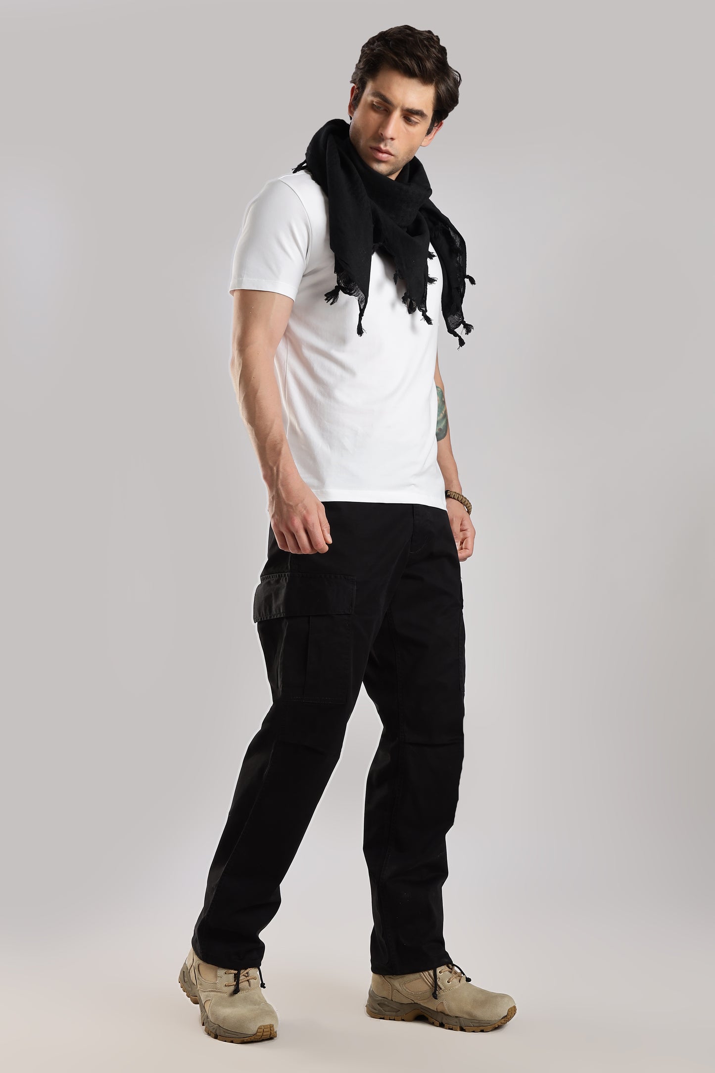 Man wearing a black tactical shemagh scarf, a white t-shirt and black cargo pants, on a gray background.