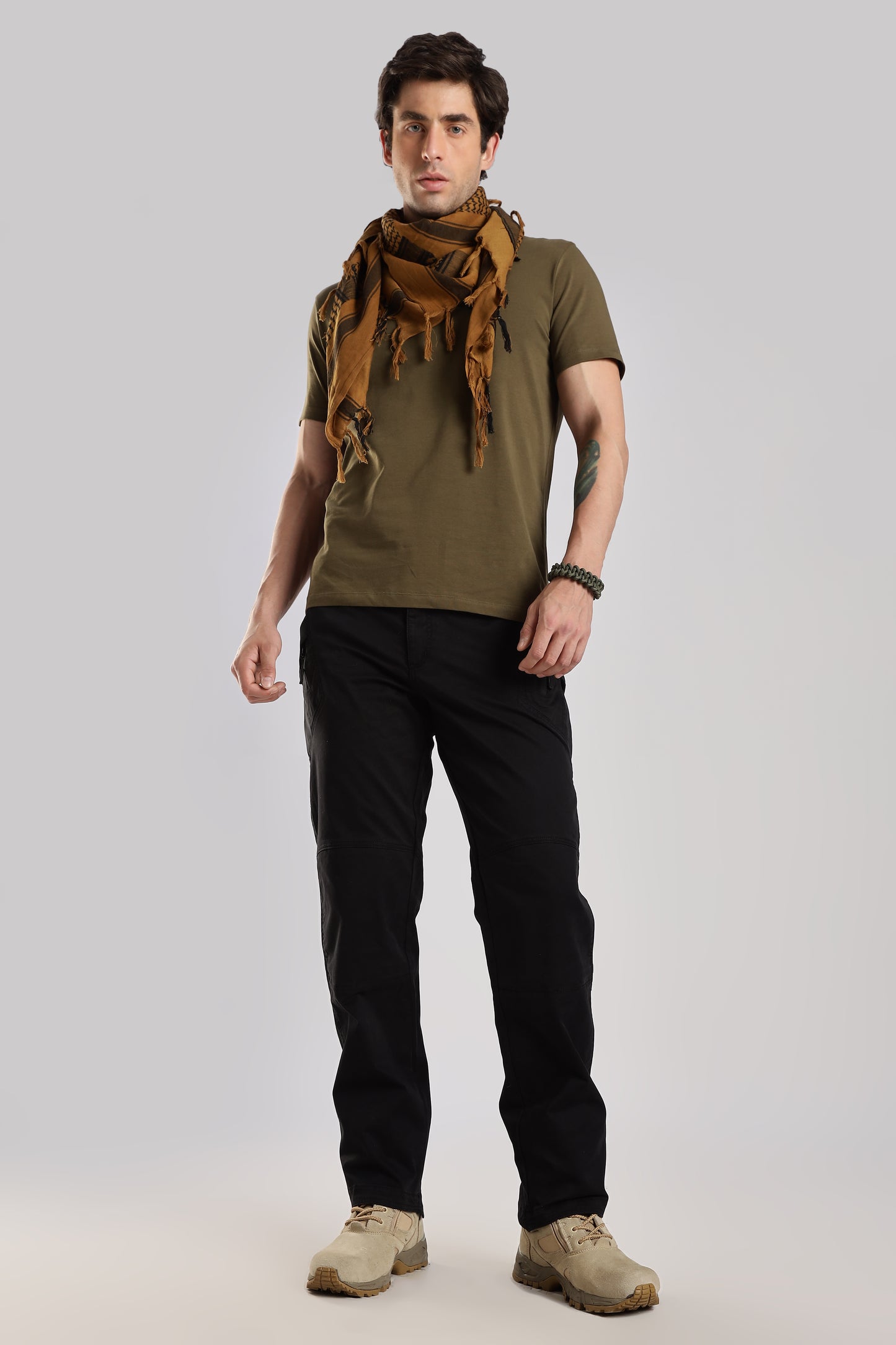 Man wearing a brown tactical shemagh scarf, an olive green tshirt and black cargo on a grey background.