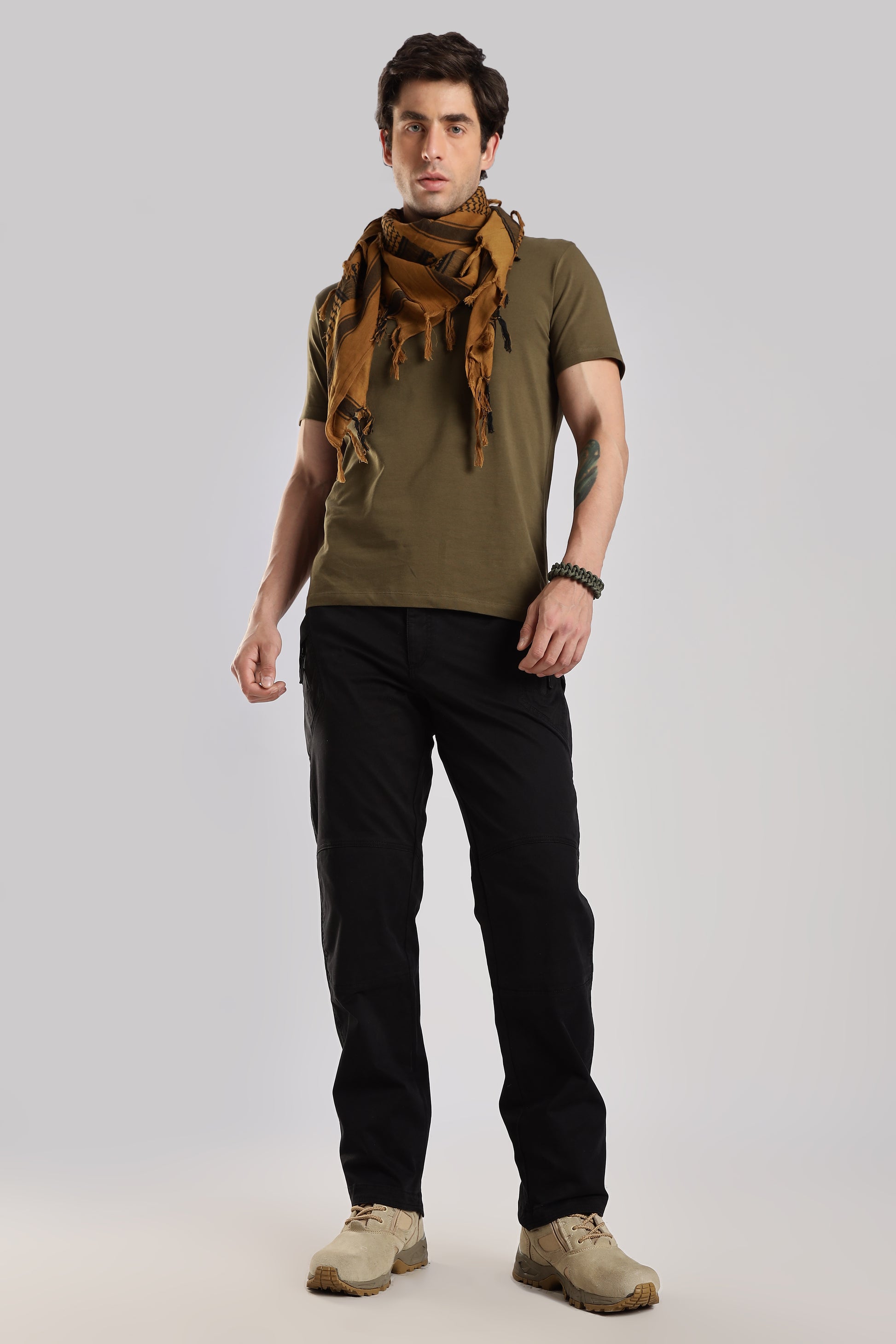 Man wearing a brown tactical shemagh scarf, an olive green tshirt and black cargo on a grey background.