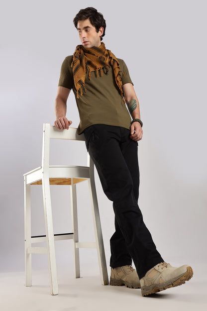 Man wearing a brown tactical shemagh scarf, an olive green tshirt, black cargo pants, and leaning against a white bar chair on a plain background.