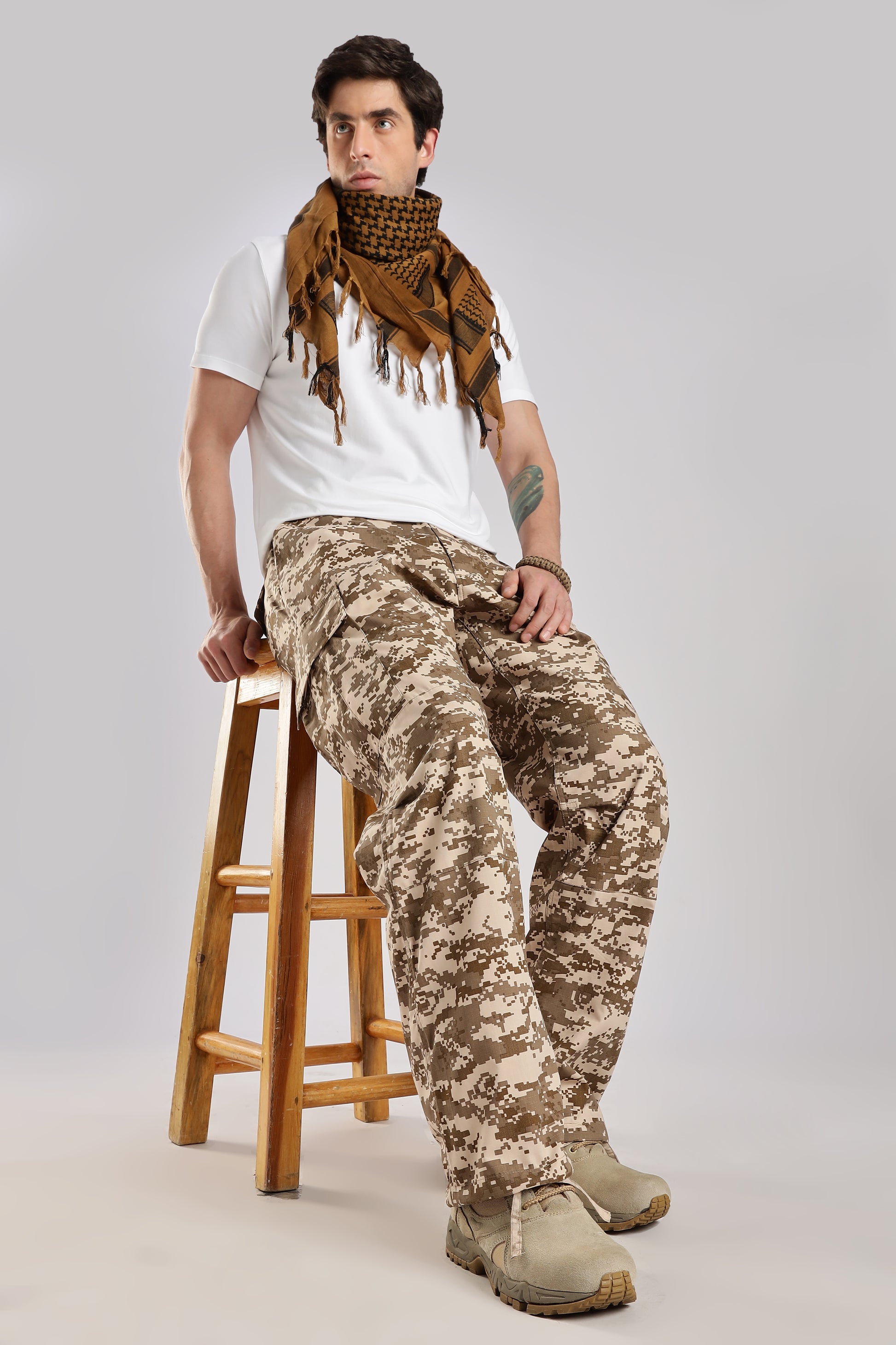 Man wearing a brown tactical shemagh, a white t-shirt and desert digital camouflage pants sitting on a wooden stool.