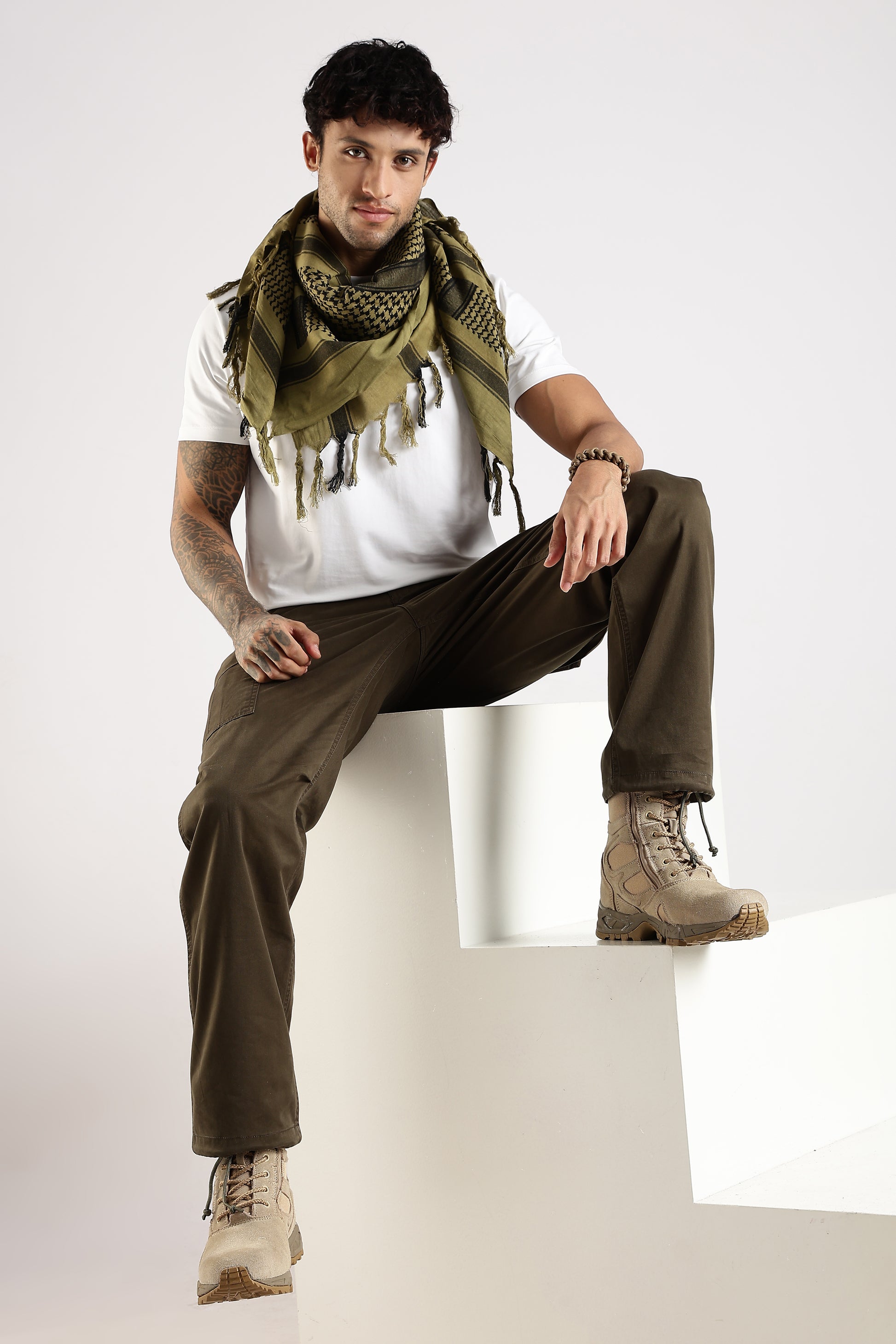 Man wearing an olive green tactical shemagh scarf, a white shirt, olive green cargo pants, and desert combat boots sitting on a white stepped block.