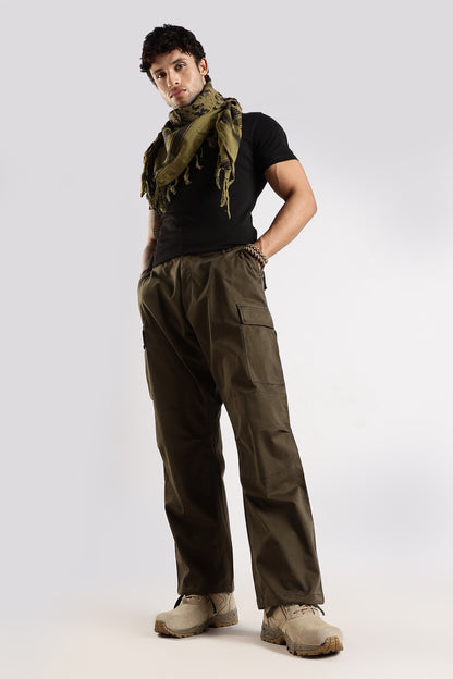 Man wearing an olive green tactical shemagh scarf in digital camo pattern, a black t-shirt, olive green paratrooper fatigue cargo pants and desert combat boots on a white background.