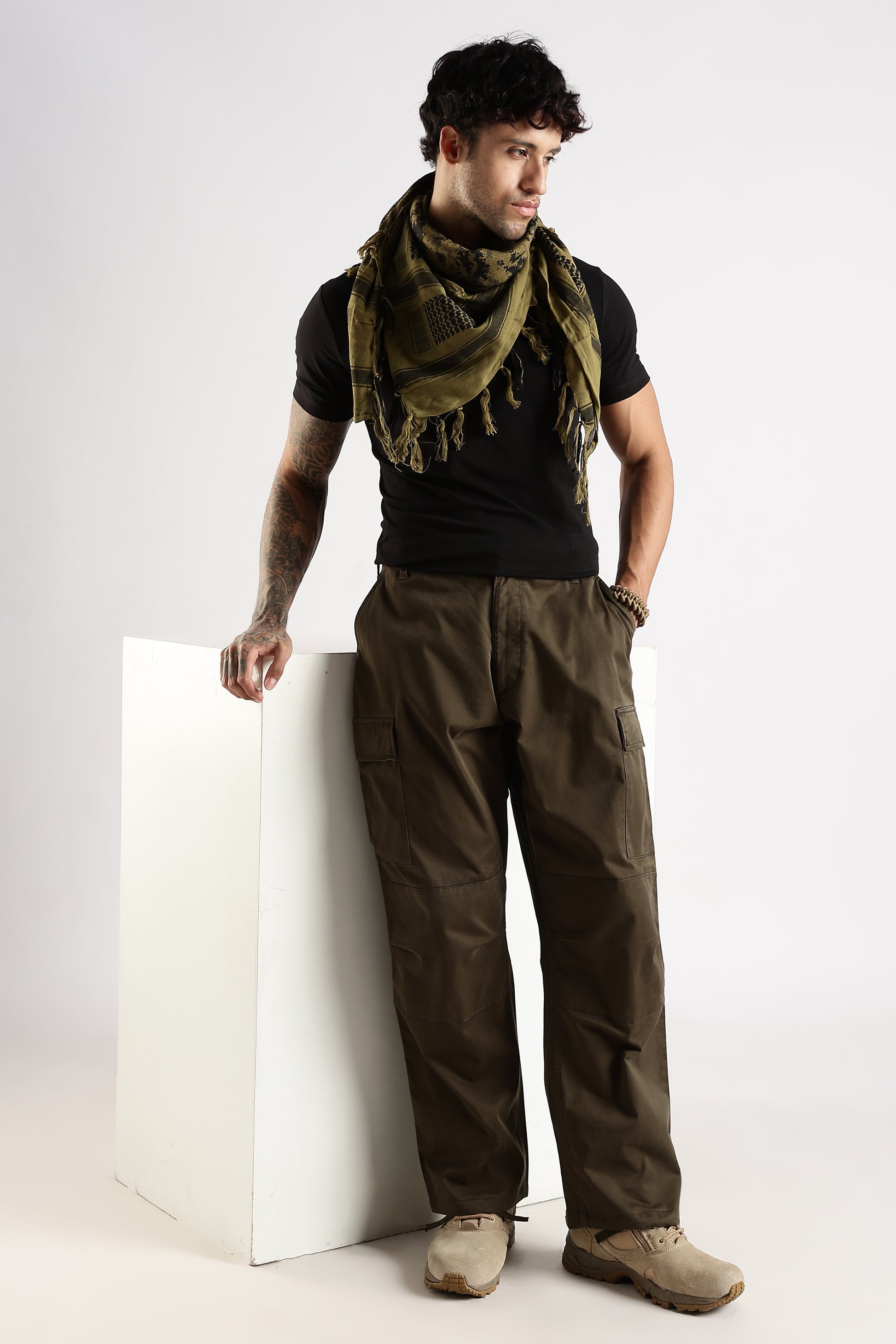 Man wearing an olive green tactical shemagh scarf in digital camouflage pattern, a black t-shirt and an olive green paratrooper cargo pants leaning against a white wooden block.